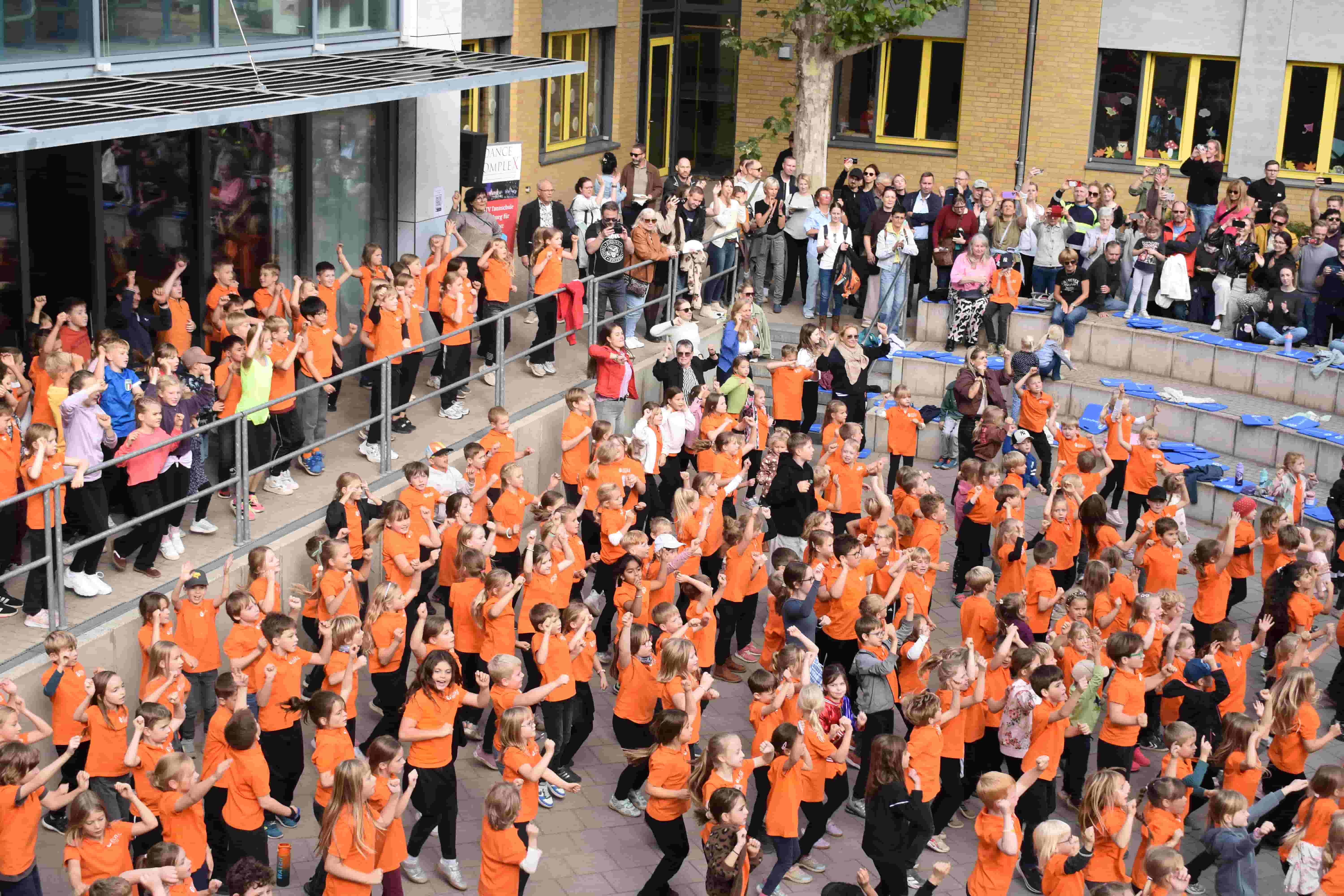 Viele Kinder in orangen Shirts tanzen im Schulhof vor zuschauenden Eltern