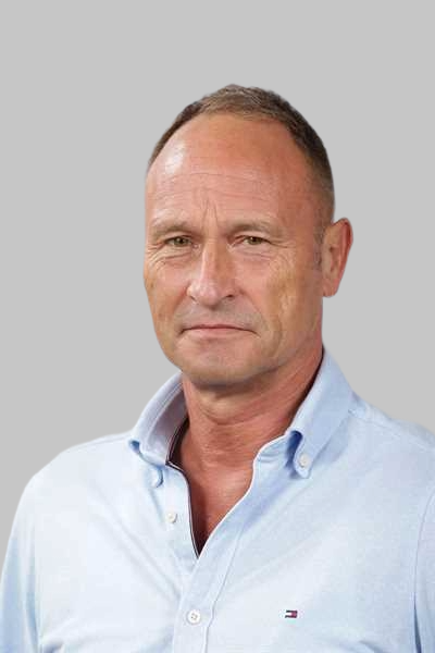 Ein Mann mit ernstem Blick traegt ein hellblaues Hemd vor grauem Hintergrund