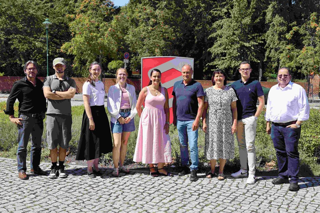 Neun laechelnde Personen stehen vor gruenen Baeumen und einem roten Schild