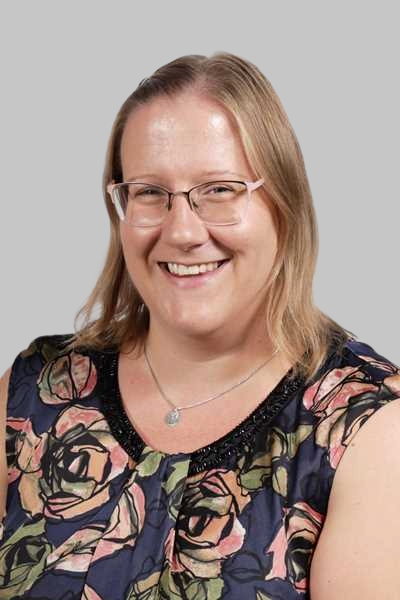 Laechelnde blonde Frau mit Brille und geblumter Bluse vor grauem Hintergrund