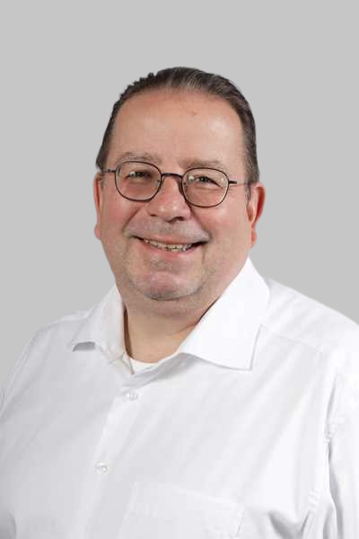 Ein laechelnder Mann mit Brille und weissem Hemd vor grauem Hintergrund
