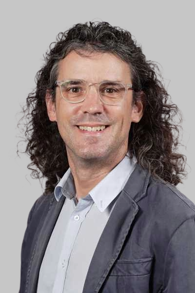 Ein laechelnder Mann mit lockigem Haar und Brille traegt ein graues Sakko