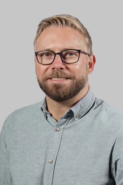 Ein laechelnder Mann mit Bart Brille und grauem Hemd vor grauem Hintergrund