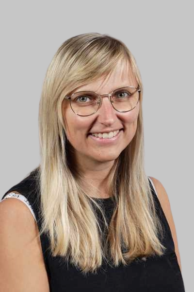 Laechelnde blonde Frau mit Brille in schwarzem Oberteil vor grauem Hintergrund