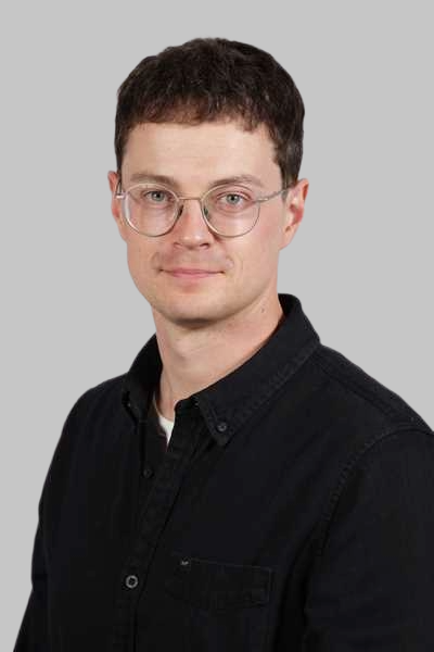 Ein laechelnder junger Mann mit Brille und schwarzem Hemd vor grauem Hintergrund