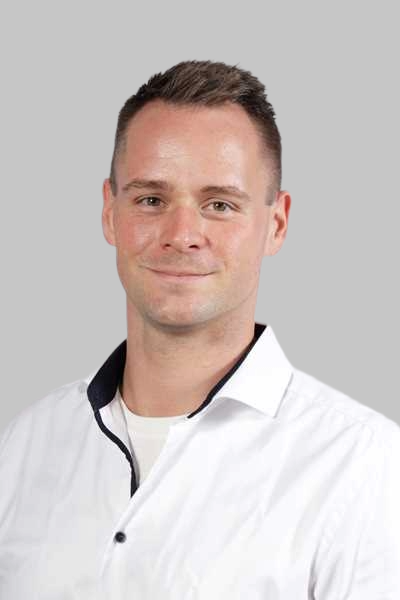 Laechelnder Mann mit kurzen braunen Haaren in weissem Hemd vor grauem Hintergrund