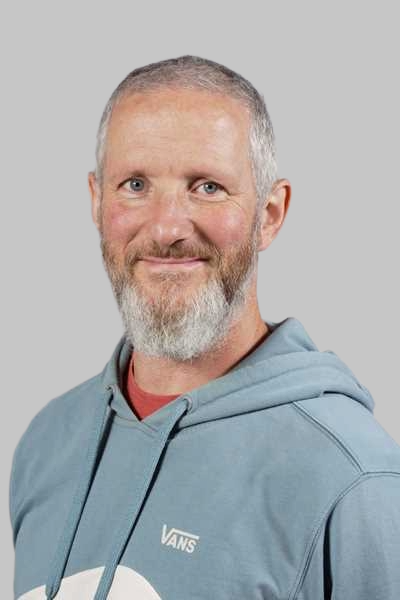 Laechelnder Mann mit grauem Bart in blauem Kapuzenpullover vor grauem Hintergrund