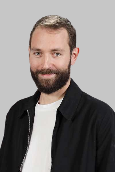 Laechelnder Mann mit Vollbart traegt schwarze Jacke und weisses Shirt