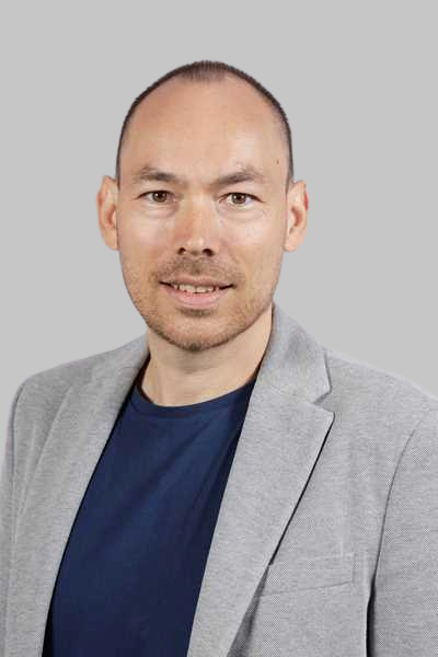 Ein laechelnder Mann im grauen Sakko und blauen Shirt vor grauem Hintergrund