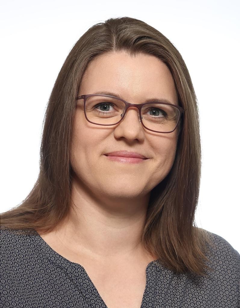 Laechelnde Frau mit brauner Brille und langem braunem Haar in grauer Bluse