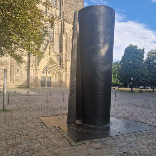 Dunkle Bronzeskulptur vor gotischer Kirche auf gepflastertem Platz bei Sonne