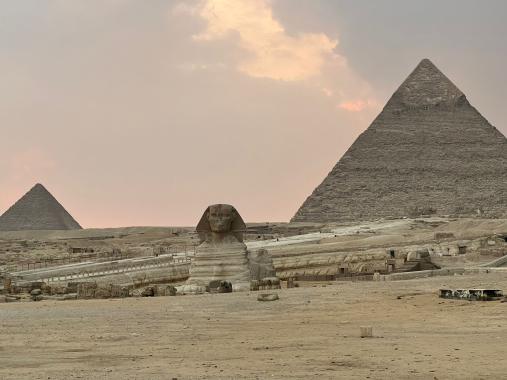 Die Sphinx von Gizeh vor zwei Pyramiden in sandfarbener Wuestenlandschaft