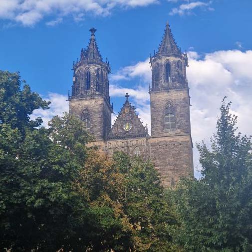 Zwei gotische Kirchtuerme aus Stein ragen hinter gruenen Baeumen empor